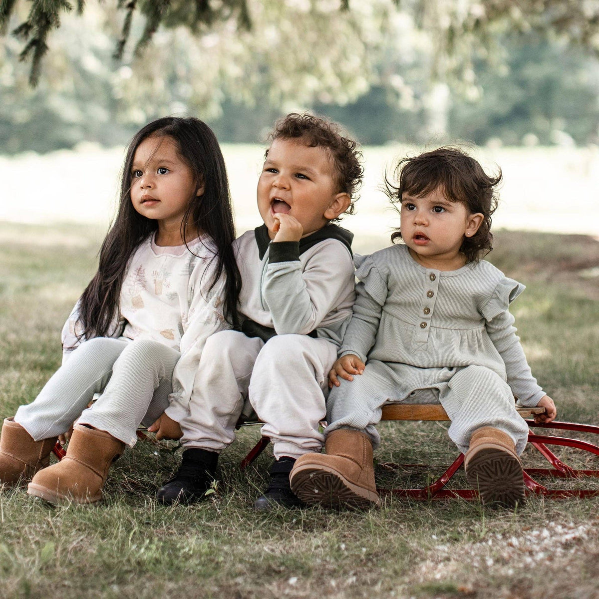 Three young children sitting together outdoors in neutral-colored outfits