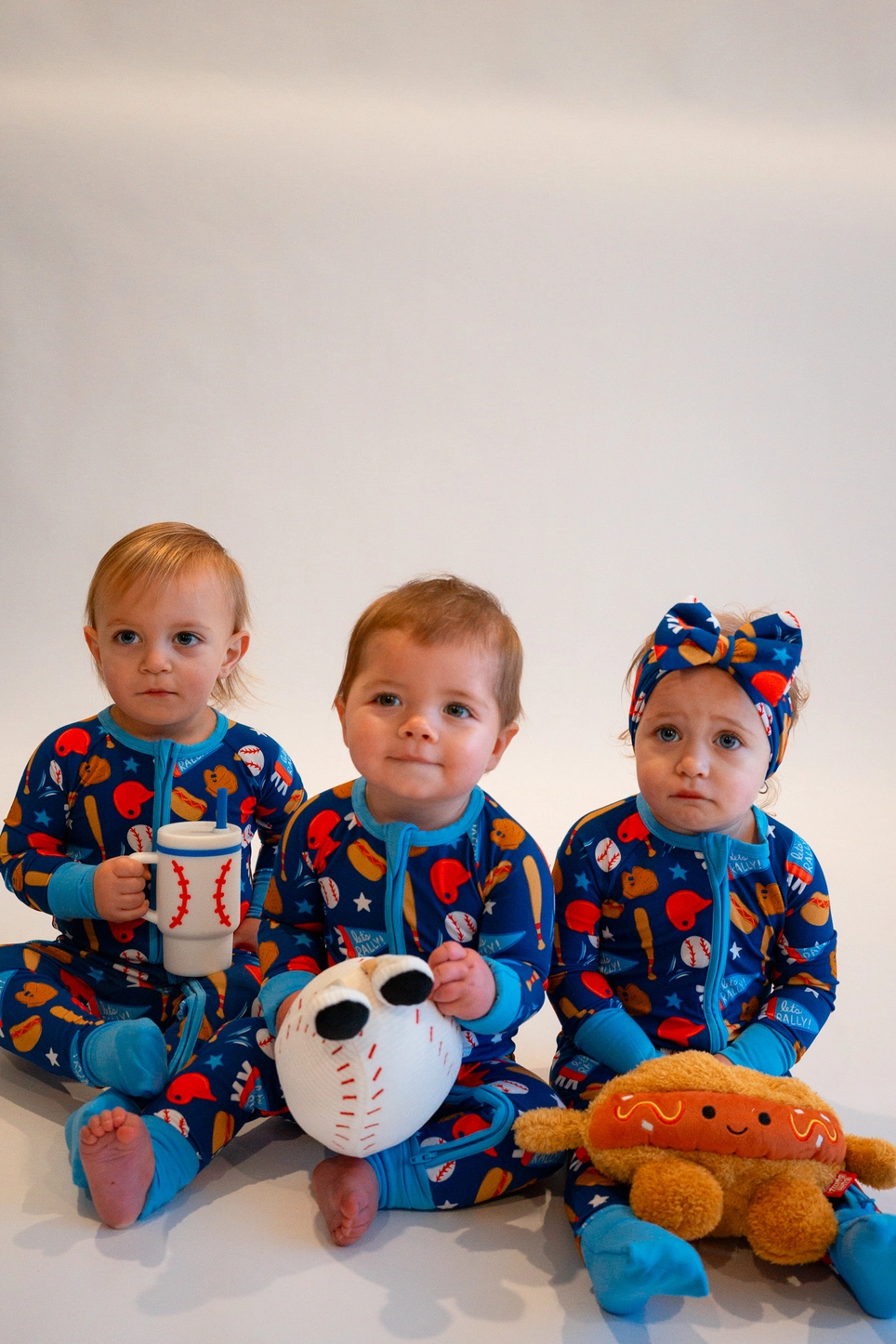 Three babies sitting together wearing matching Baseball Bamboo Zippy Pajamas with baseball plush toys