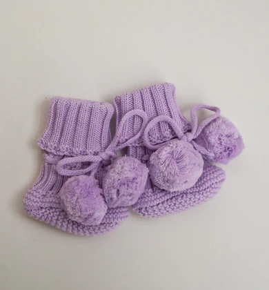 Pompom knitted baby booties in orchid purple, showing oversized pompom accents and drawstring closure