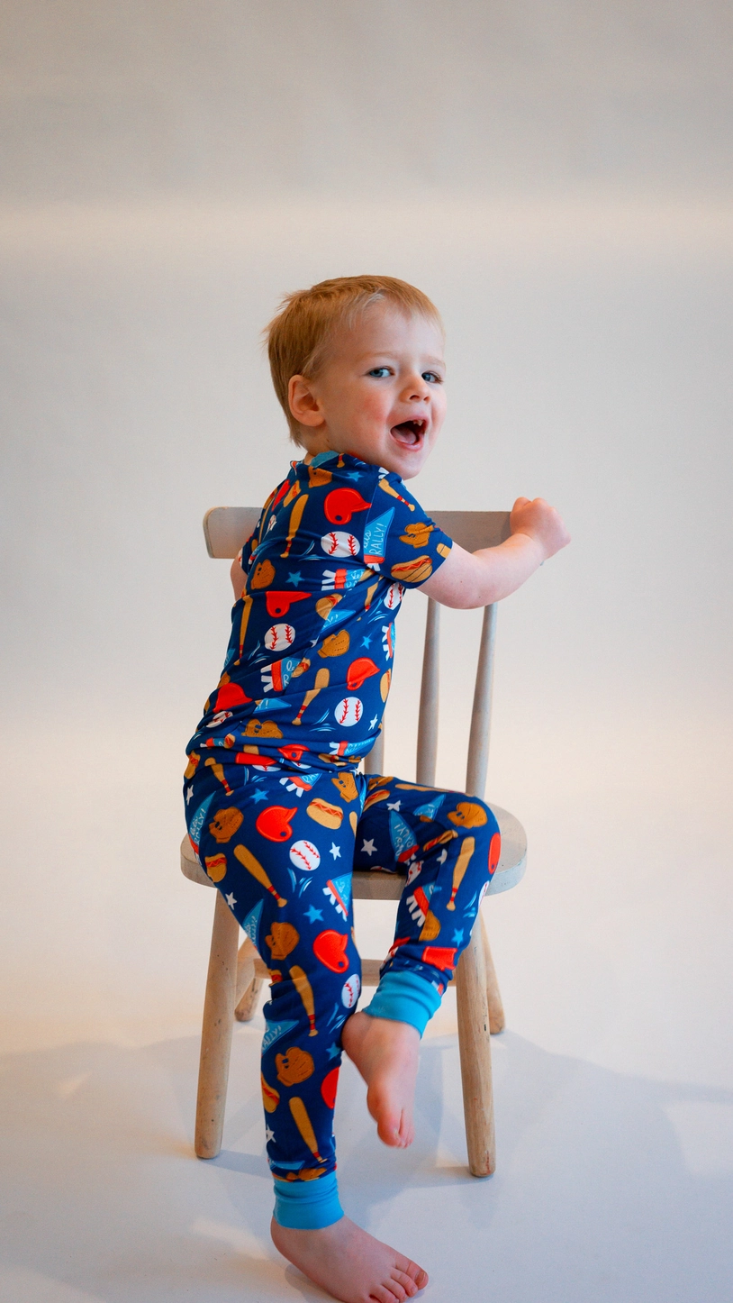Toddler sitting on chair wearing Baseball Bamboo Two-Piece Pajamas with baseball gloves bats and hotdog print