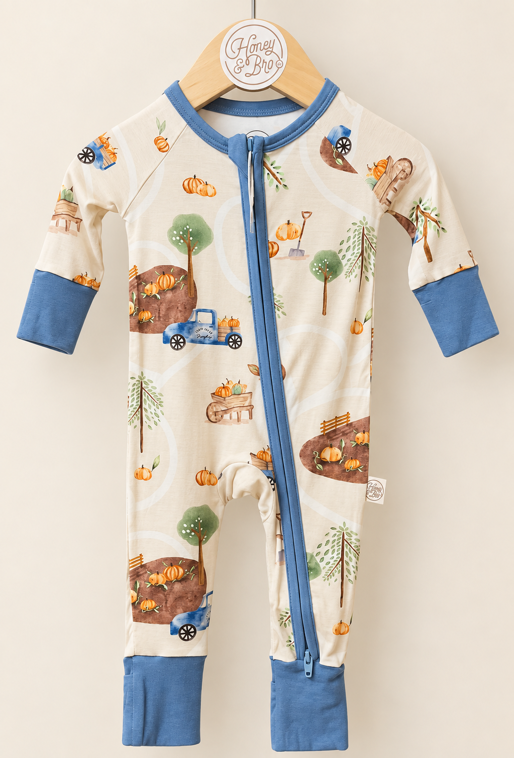 Harvest Haul Zinza zippy on a hanger: cream autumn print with pumpkins, trees and trucks, and blue trim.