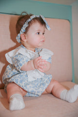 Baby girl sitting in blue floral lace romper with matching headband - side profile on pink chair