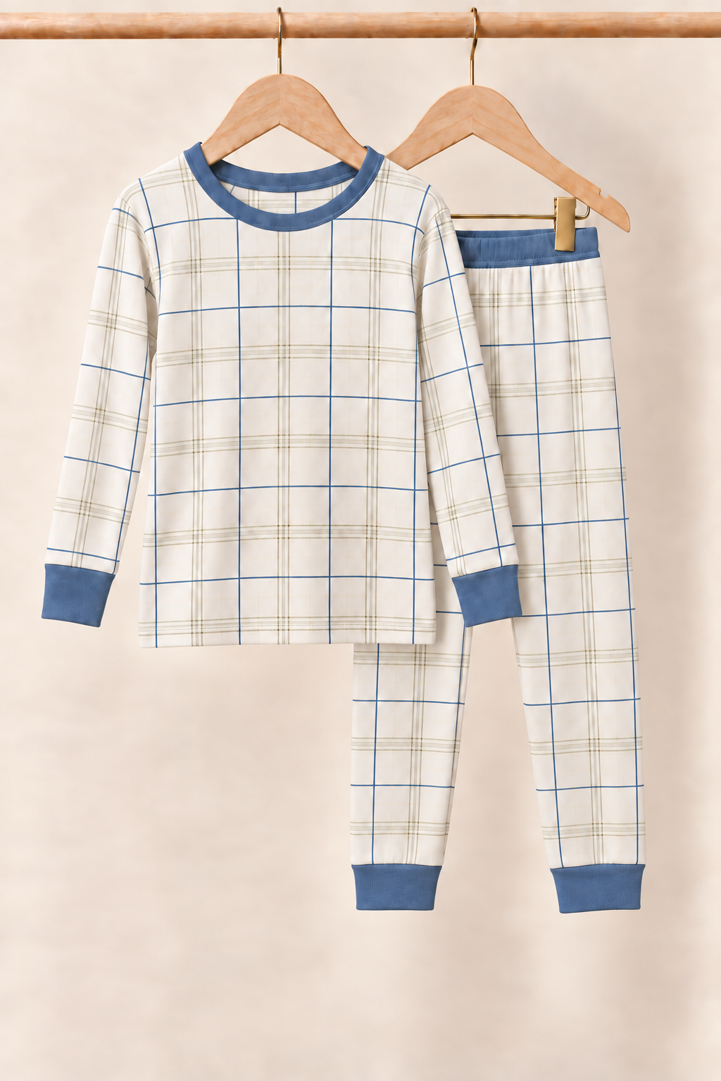Fireside Windowpane ivory plaid kids two-piece pajamas with slate blue neckline, cuffs and pants waistband on wooden hangers against a creamy background